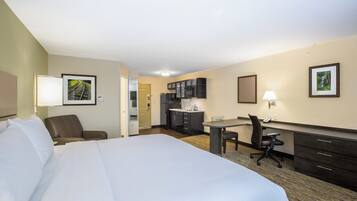Habitación de hotel con una cama grande, un escritorio, una silla, una kitchenette y dos cuadros en las paredes.
