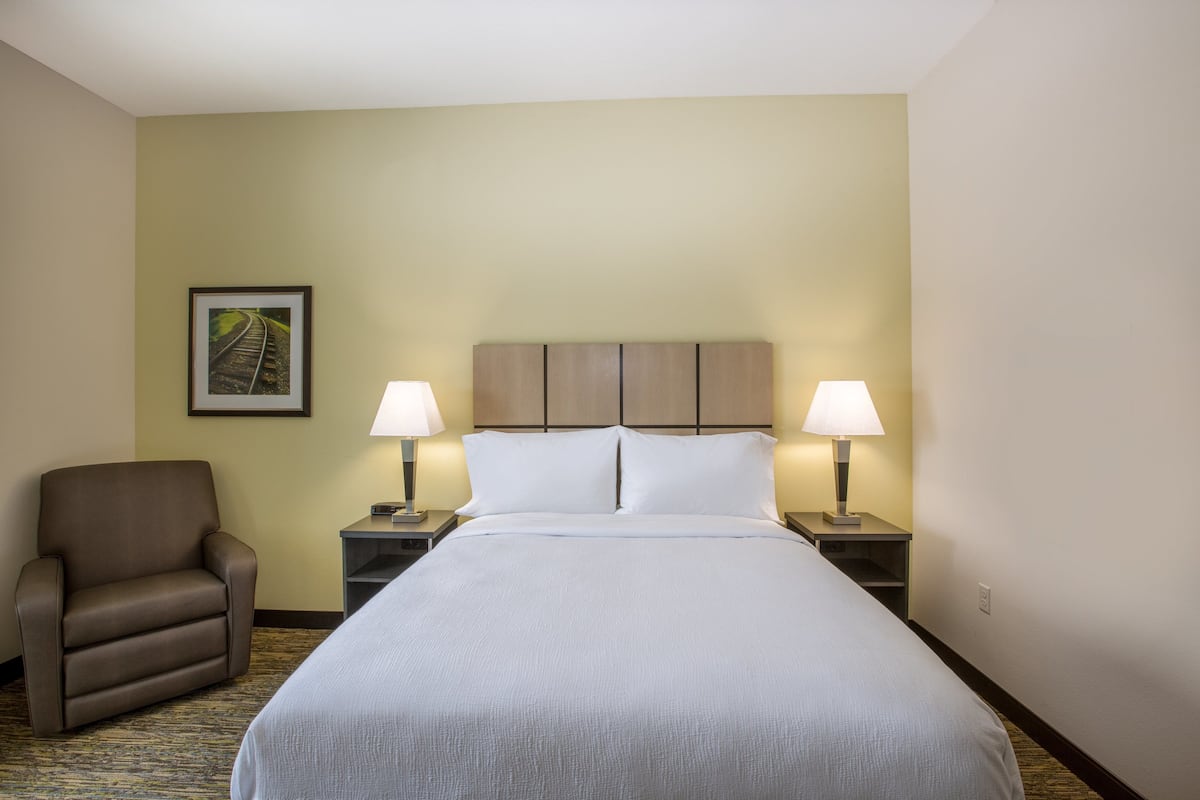 A hotel room with a large bed, two bedside tables with lamps, a brown armchair, and a framed picture on the wall.