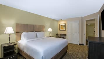 A hotel room with a large bed, two bedside tables with lamps, a desk with a chair, a wardrobe, and a door leading to another room.
