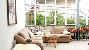 A living room with a beige sectional sofa, a small round wicker table, and a large window with a view of a garden and a brick wall. - Lush & Co Auckland Bed & Breakfast (Auckland)