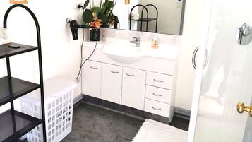 A bathroom with a white sink, mirror, and a black hairdryer mounted on the wall. There are white cabinets below the sink and a black shelf to the left.