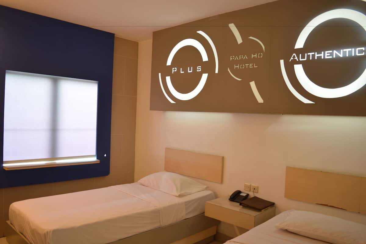 A hotel room with two beds, a window with blinds, a wall-mounted phone, and a sign indicating 'Plus' and 'Authentic' hotel branding.