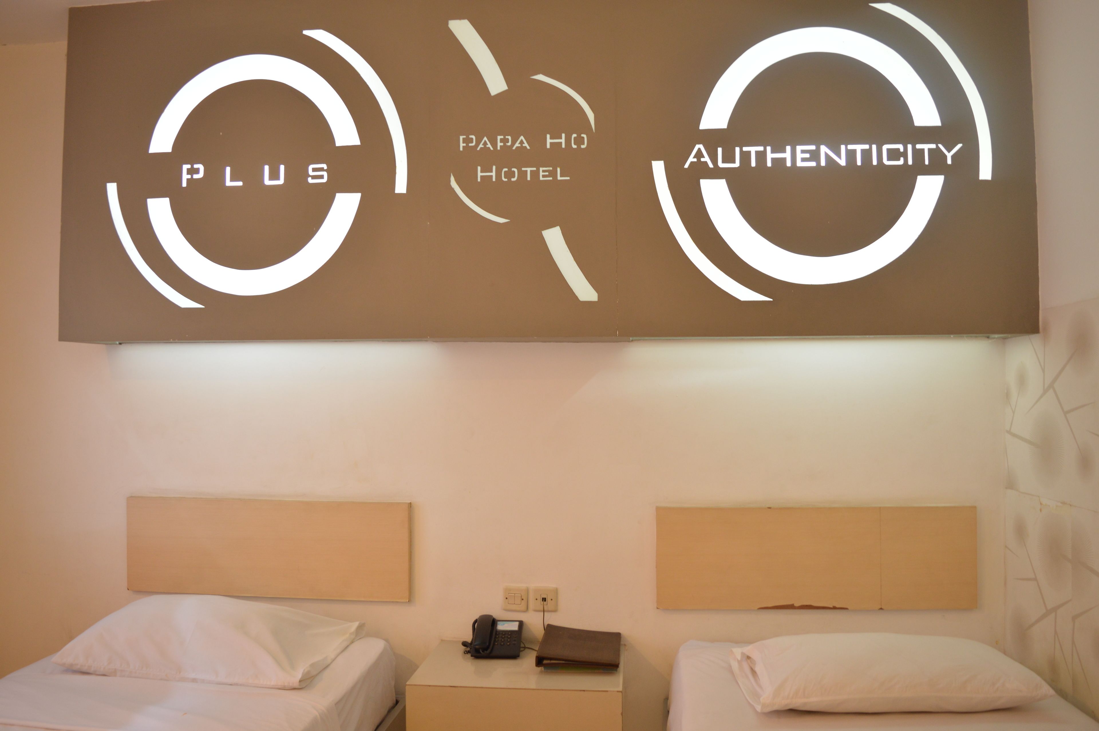 Et hotelværelse med to senge, et lille natbord med telefon og et skilte på væggen med hotellets navn og logo.