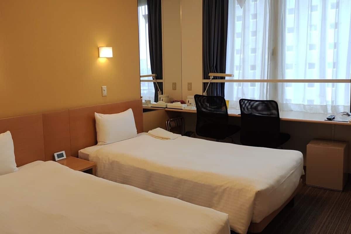 Ein Hotelzimmer mit zwei Betten, einem Schreibtisch, einem Stuhl und einem Vorhangfenster.