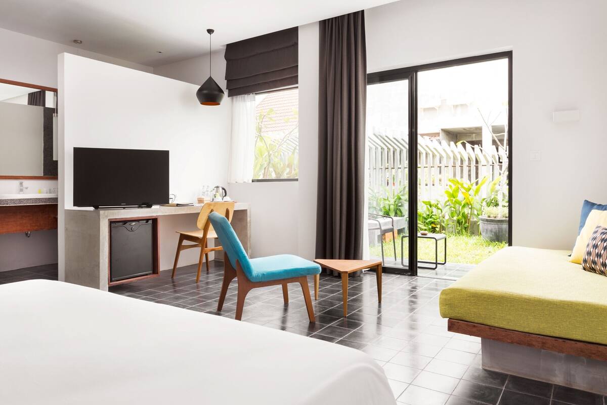 A modern hotel room with a bed, a desk with a TV, a chair, and a view of the outdoors.