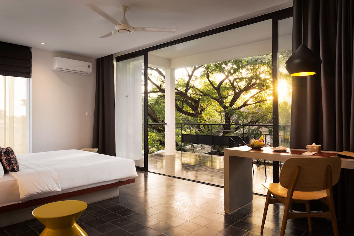 A modern hotel room with a bed, a desk, a chair, and a balcony with a view of trees.