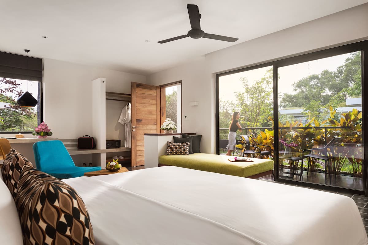 A modern hotel room with a large bed, a balcony with plants, a desk, and a wardrobe.