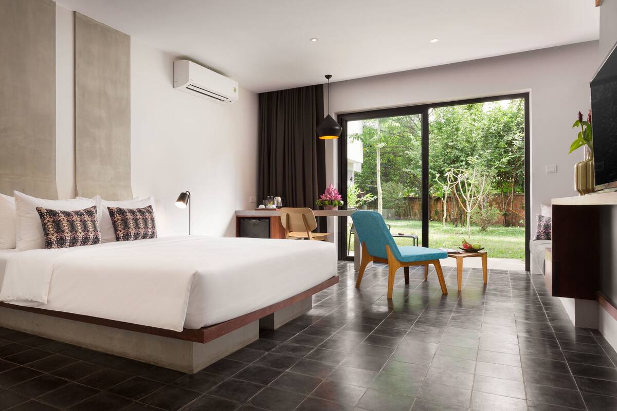A modern hotel room with a large bed, a desk, a chair, and a view of the garden.
