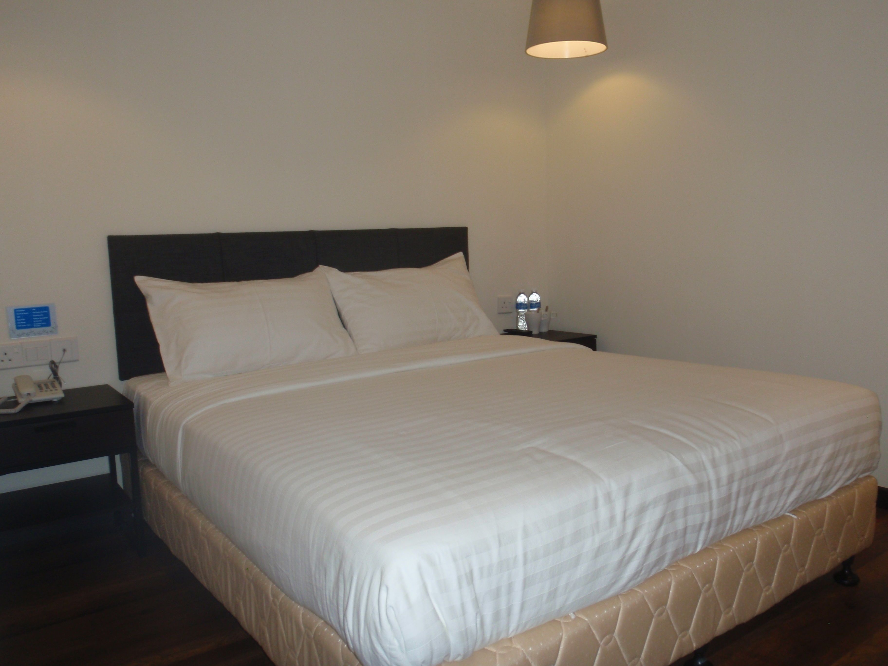 Una cama bien hecha con cabecera, una mesita de noche con lámpara y un enchufe de pared.
