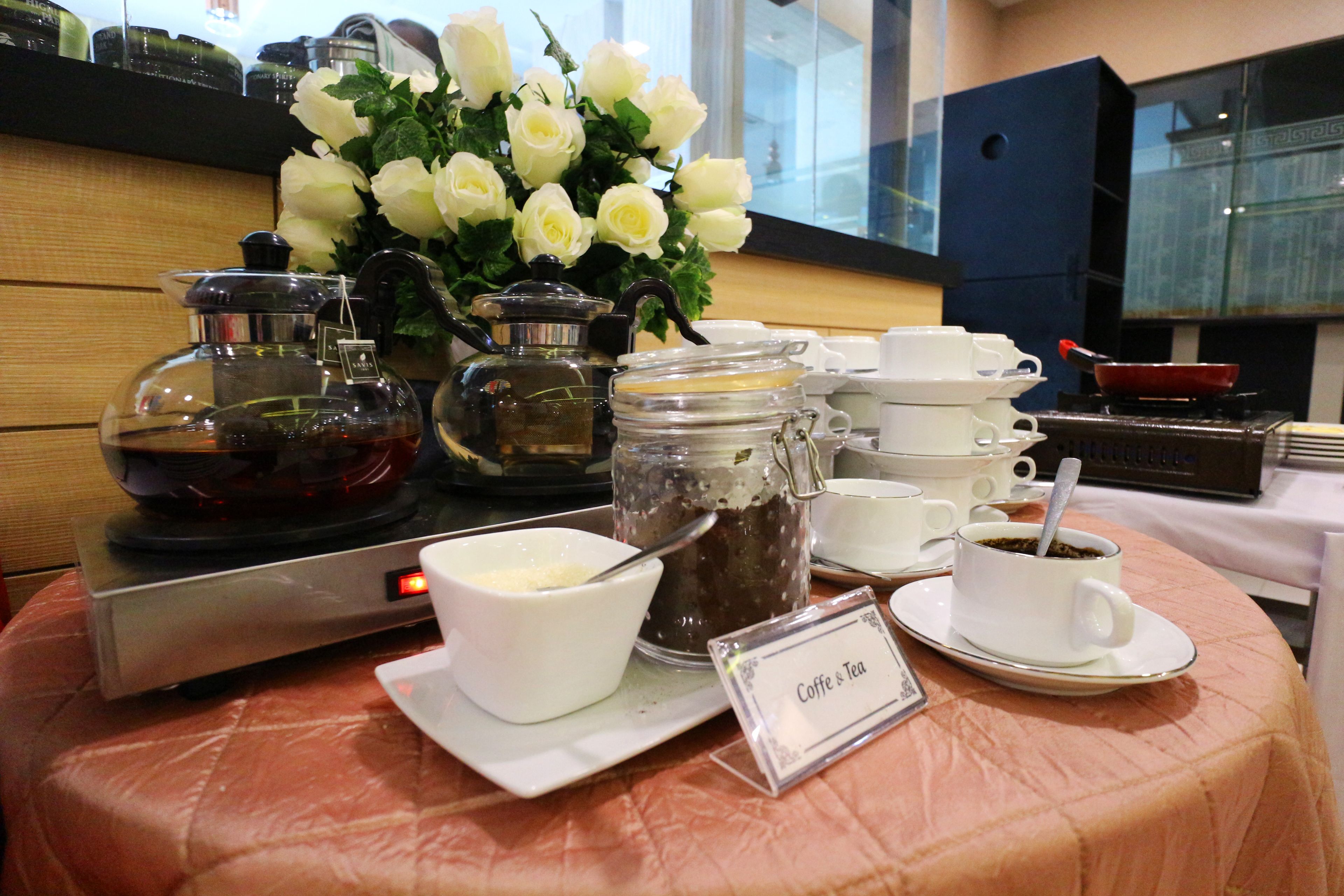 Een buffettafel met koffie en thee, een koffiezetapparaat en een vaas witte bloemen.