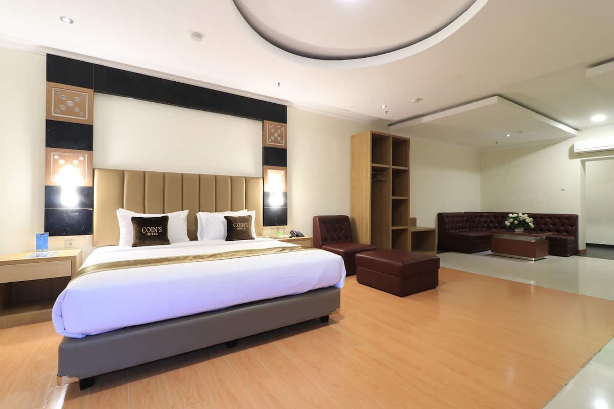 A modern hotel room with a large bed, a seating area with a sofa and armchairs, a wooden cabinet, and a large mirror.