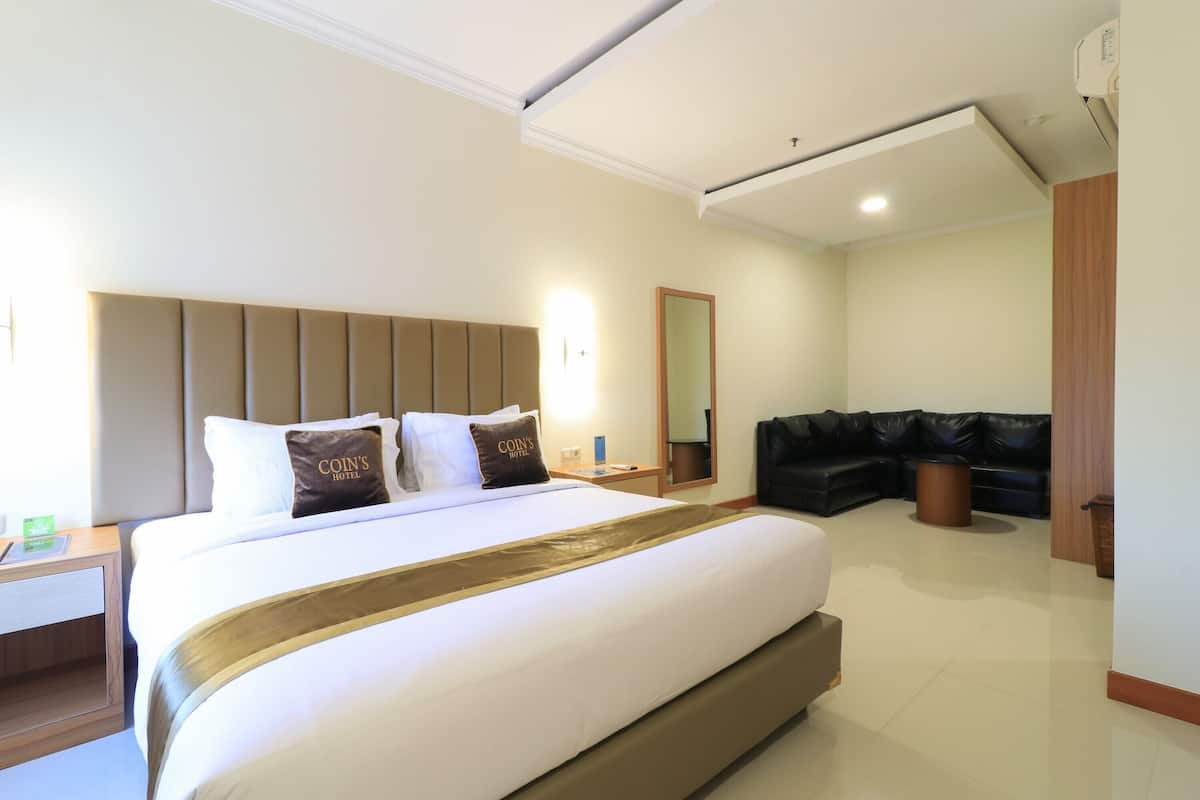 A hotel room with a large bed, a headboard, a bedside table, a mirror, a black leather sofa, and a trash bin.