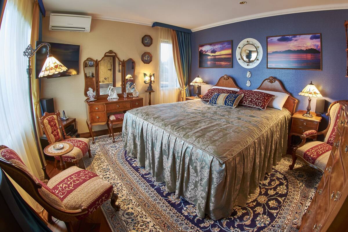Un espacioso dormitorio con una cama grande, sillas ornamentadas, un aparador de madera y un candelabro clásico.