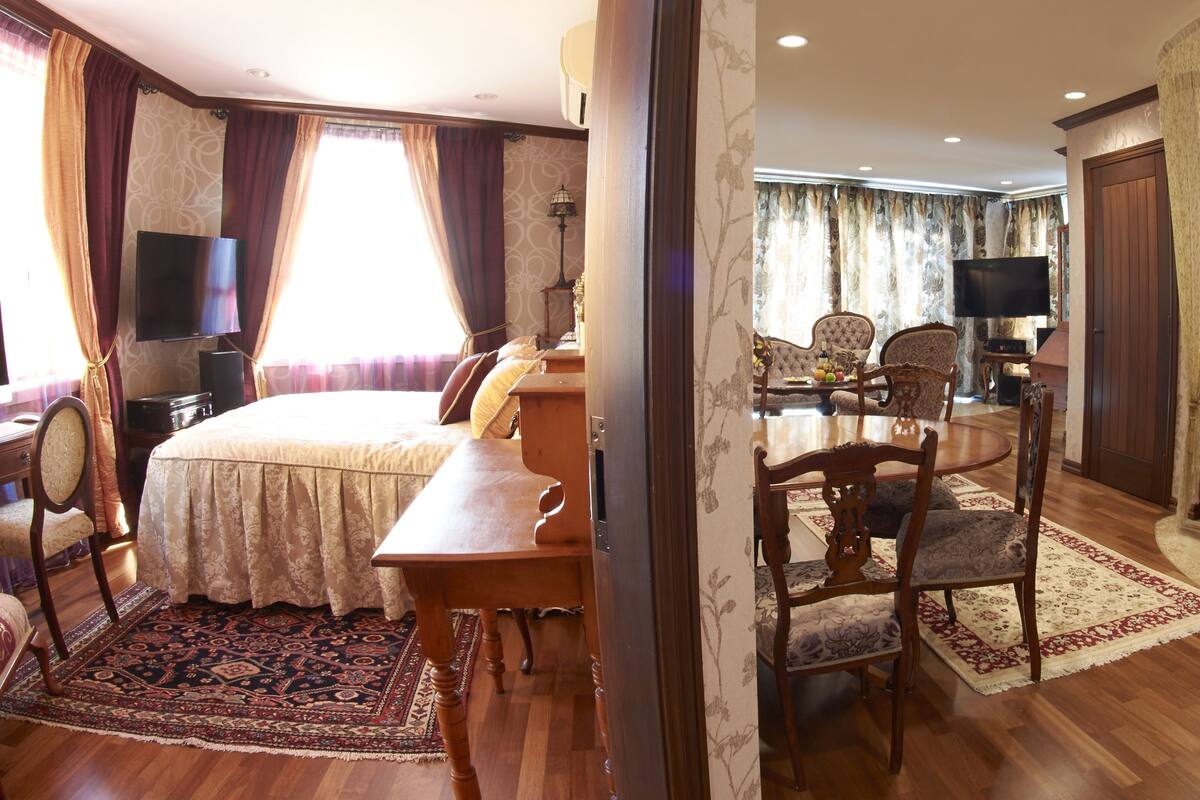 Habitación de hotel con cama, escritorio, mesa de comedor con sillas, televisor y baño con lavamanos y espejo.