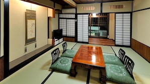 Une pièce avec un sol recouvert de tatamis, une table et des chaises en bois, des portes coulissantes et une œuvre d’art traditionnelle japonaise accrochée au mur.