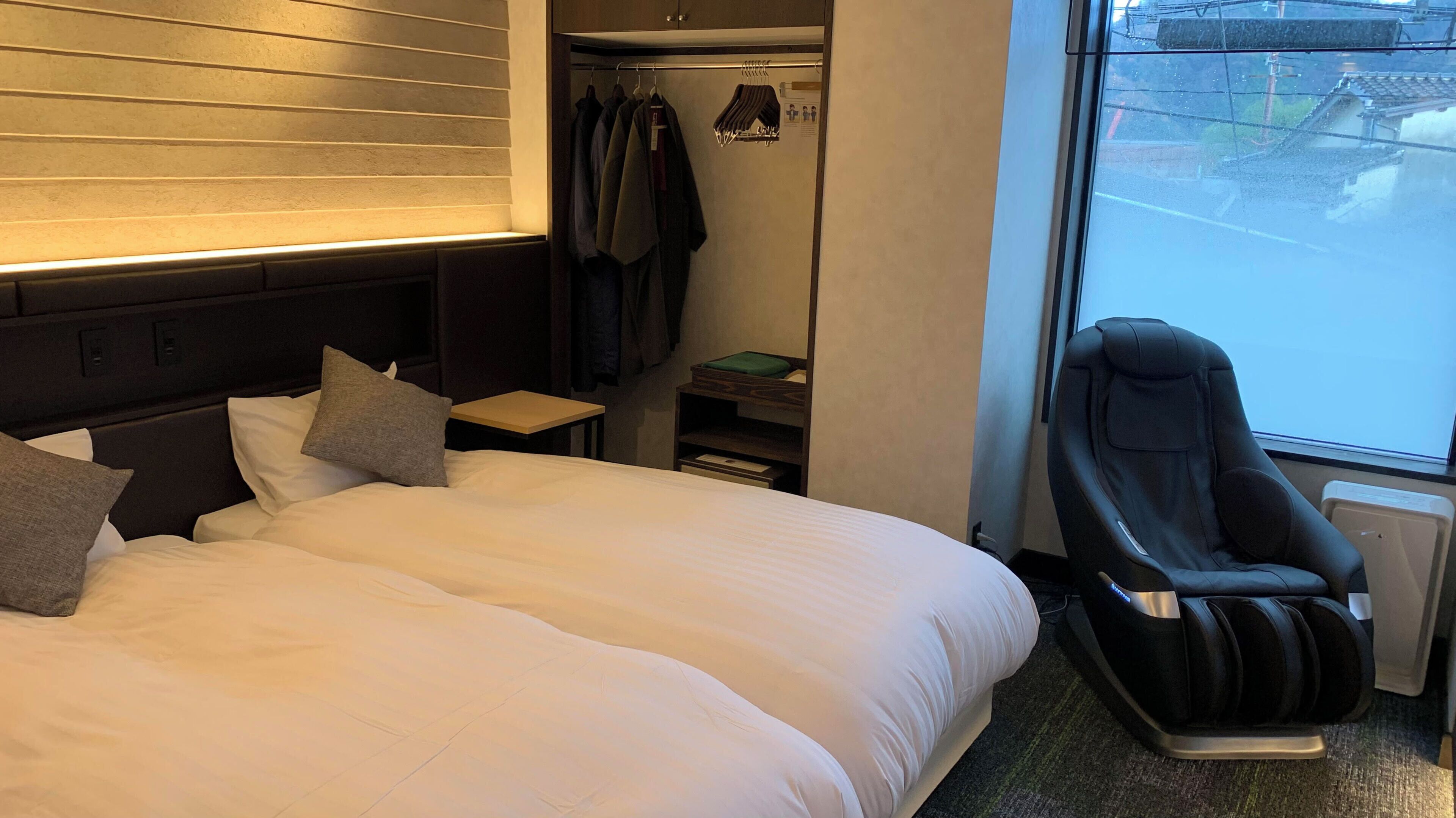 Une chambre d’hôtel avec un lit, un fauteuil massant, une armoire avec des vêtements suspendus et une fenêtre.