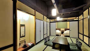Une pièce traditionnelle avec un sol recouvert de tatamis, une petite table et des coussins, des portes coulissantes en bois et un luminaire au plafond.