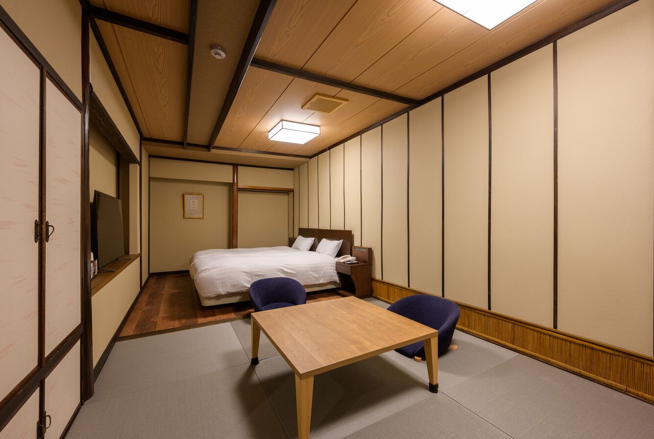 A traditional Japanese-style room with tatami flooring, a bed with white bedding, a wooden table, and two blue chairs.