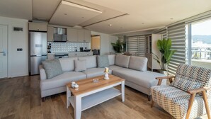A modern living room with a sectional sofa, a wooden coffee table, and a patterned armchair. A kitchen with white cabinets and a stainless steel refrigerator is visible in the background. - Aurasia SeaSide Hotel (Marmaris)