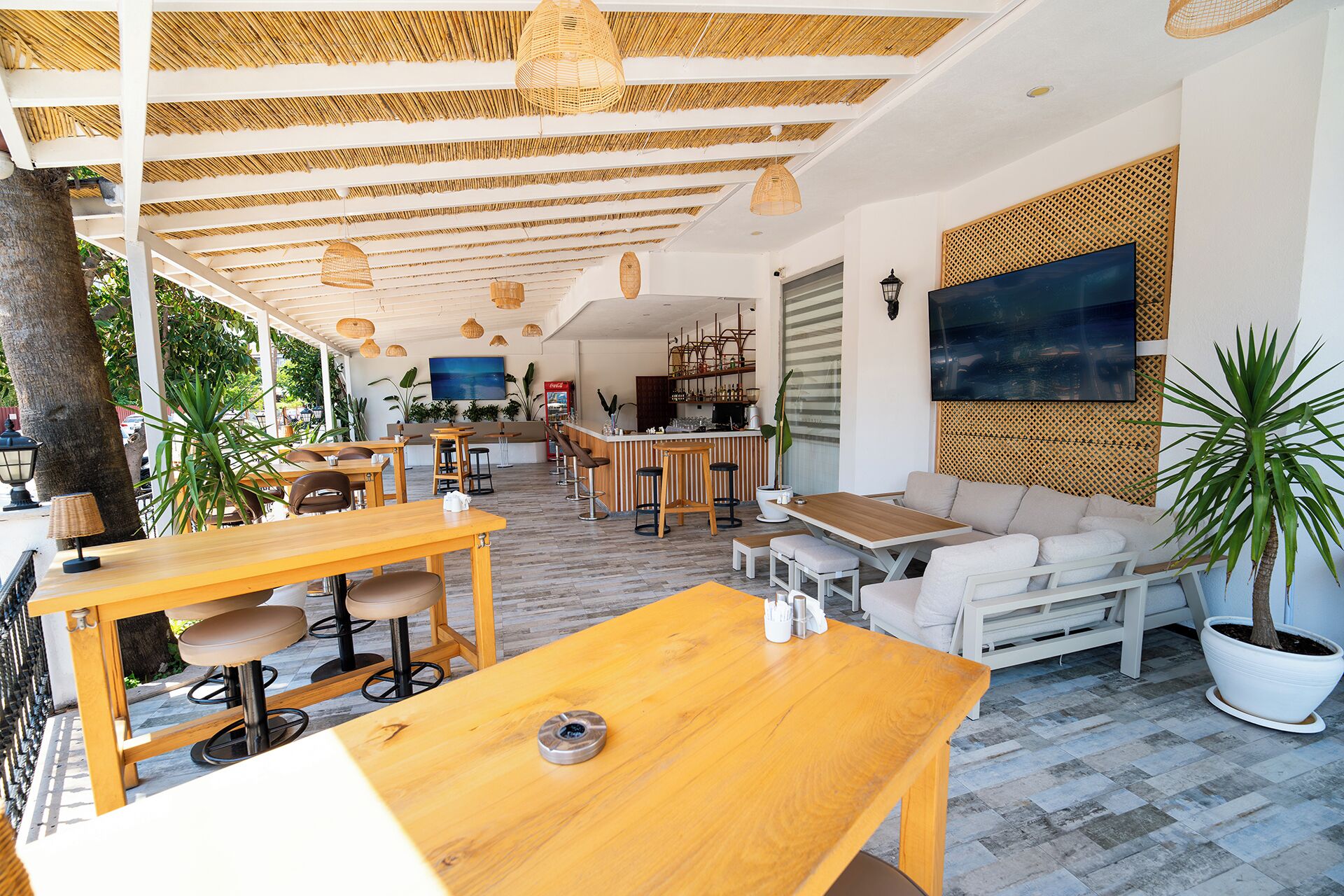 Un área de comedor exterior moderna con mesas y sillas de madera, una barra y un espacio de descanso con un sofá y una planta grande en maceta.