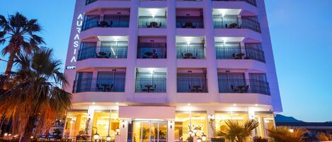 A multi-story hotel with balconies, a well-lit entrance, and potted plants around the building.