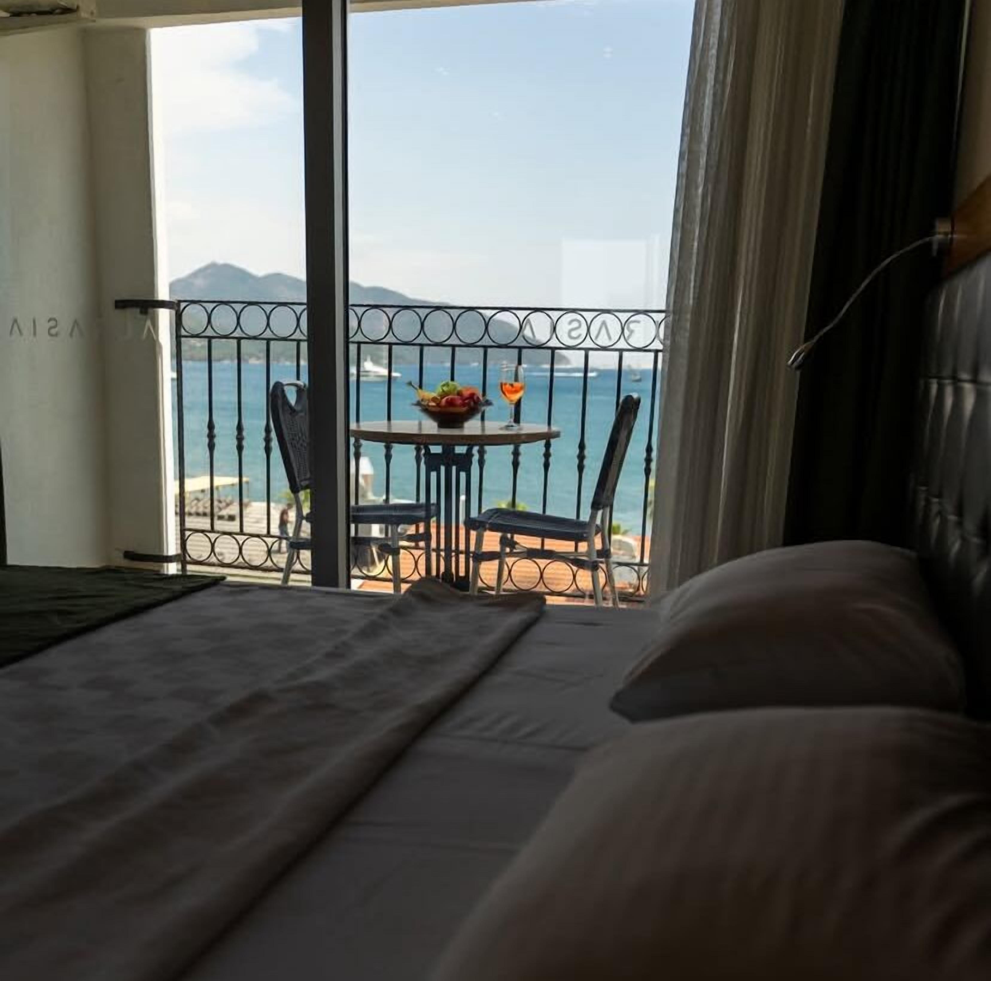 A hotel room with a bed, a small table with a fruit bowl and a glass of wine, and a view of the sea and mountains.