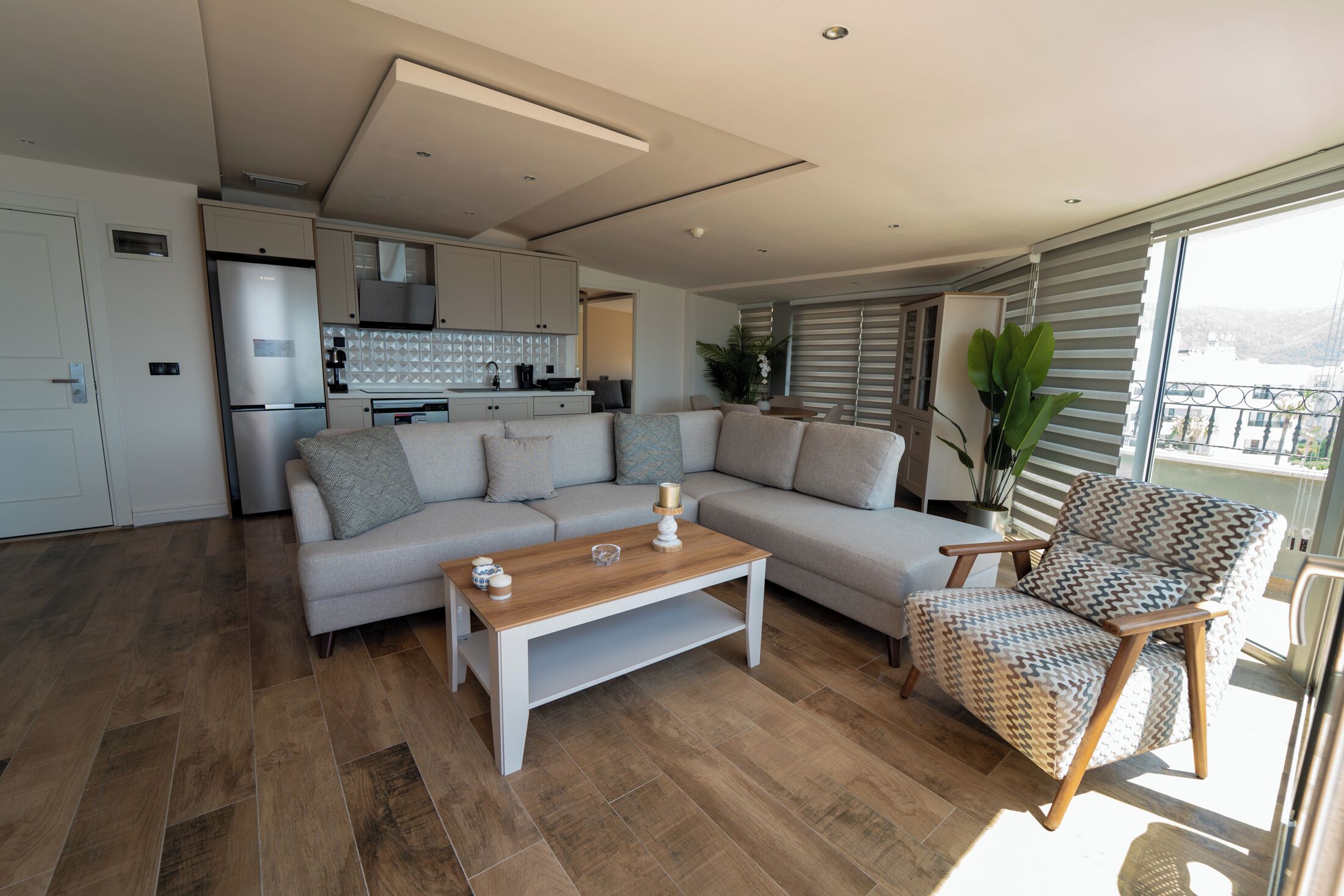 A modern living room with a sofa, a wooden coffee table, and a patterned armchair. A kitchen with white cabinets and a refrigerator is visible in the background.
