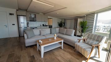 Una sala de estar moderna con un sofá, una mesa de centro de madera y un sillón estampado. Se ve una cocina con armarios blancos y un refrigerador al fondo.