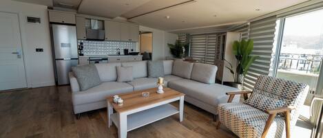 A modern living room with a sofa, a wooden coffee table, and a patterned armchair. A kitchen with white cabinets and a refrigerator is visible in the background.