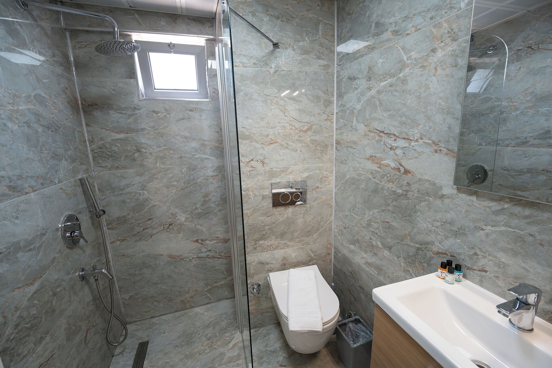 Un baño con ducha envuelta en vidrio, inodoro, lavamanos con espejo y un cesto de basura.