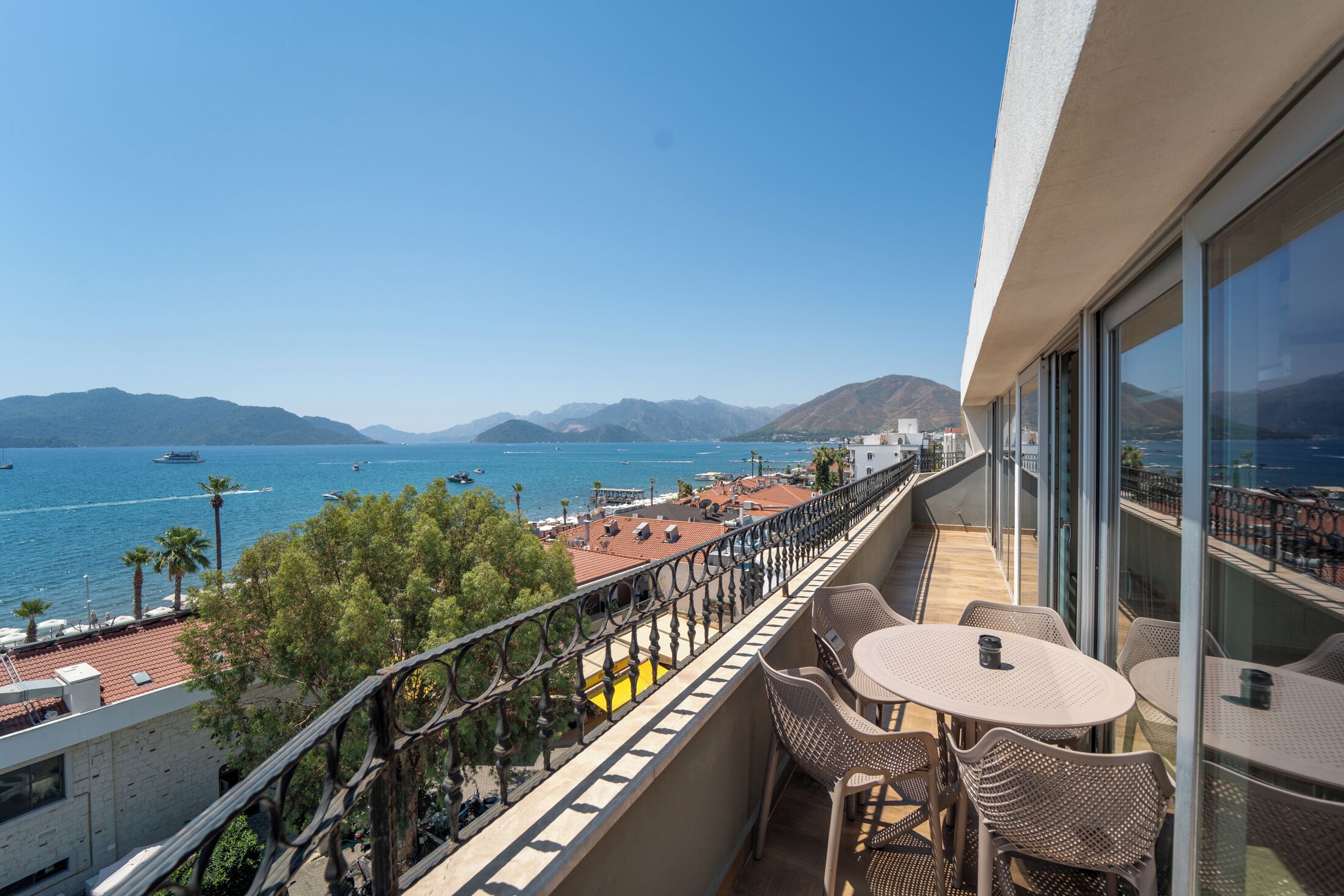 A balcony with outdoor seating overlooking a coastal area with boats and mountains.