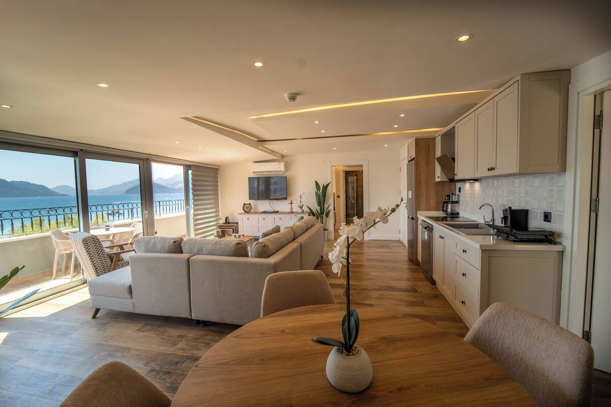 A modern living room with a large sofa, a dining table with a vase of flowers, and a kitchen area with white cabinets and a view of the sea.