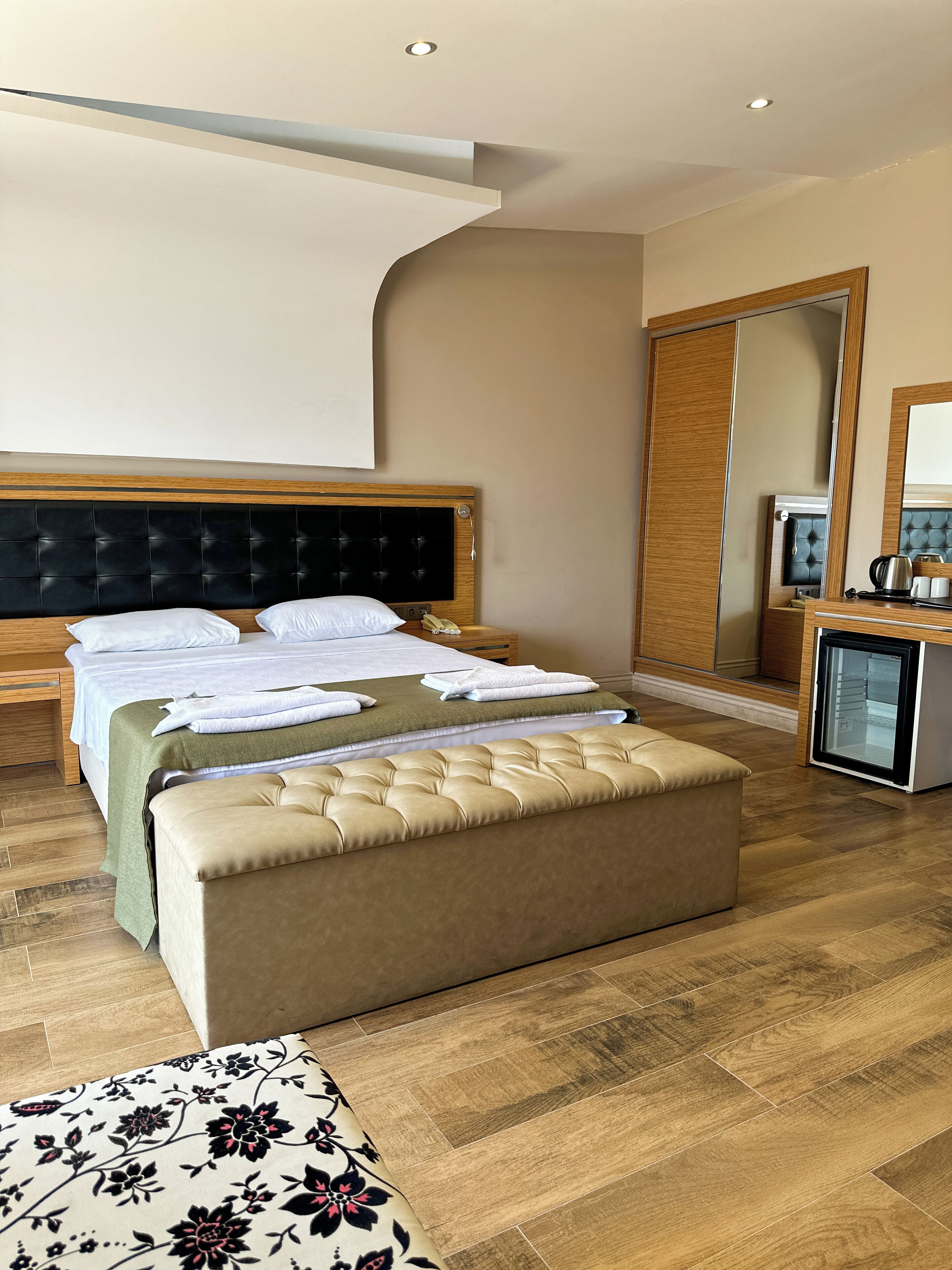 A modern hotel room with a large bed, a small seating area, a minibar, and a mirror.