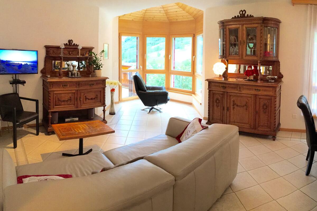 Ein Wohnzimmer mit einer Couch, einem Esstisch mit Stühlen, einem Fernseher, einem Schrank und einem großen Fenster mit Vorhängen.