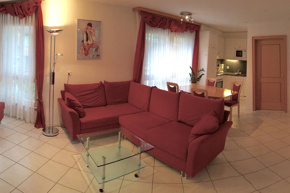 Ein Wohnzimmer mit rotem Sofa, einem Glastisch, einem Esstisch mit Stühlen, einer Küchenzeile und einem an der Wand befestigten Fernseher.