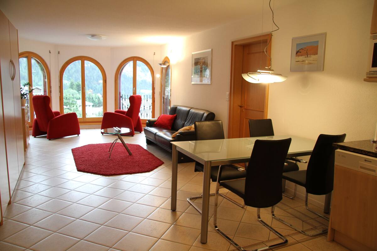 Ein geräumiger Wohnbereich mit einem Glastisch, roten Stühlen, einem schwarzen Ledersofa und einem roten Teppich. Große Fenster mit Rundbögen, eine Holztür und eine wandmontierte Leuchte sind ebenfalls vorhanden.