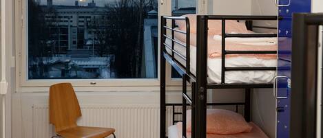 A dormitory room with bunk beds, a chair, a window with a city view, and a radiator.