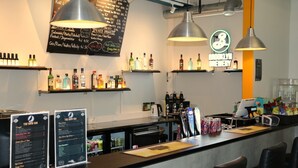 Una zona de bar con un panel de menú, estantes con botellas y una barra con grifos y vasos.