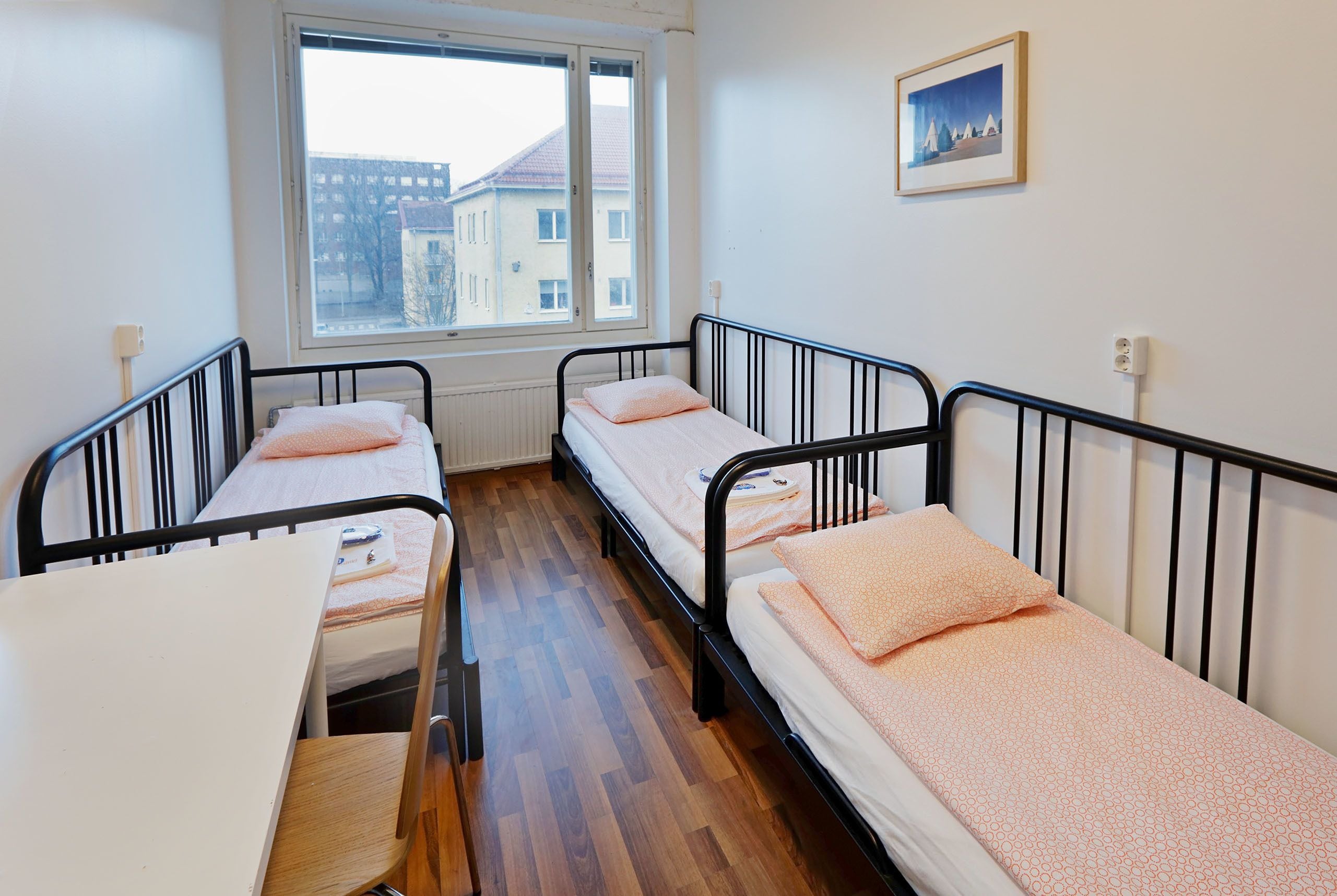 A dormitory room with three single beds, a wooden floor, a window with a view of buildings, and a framed picture on the wall.