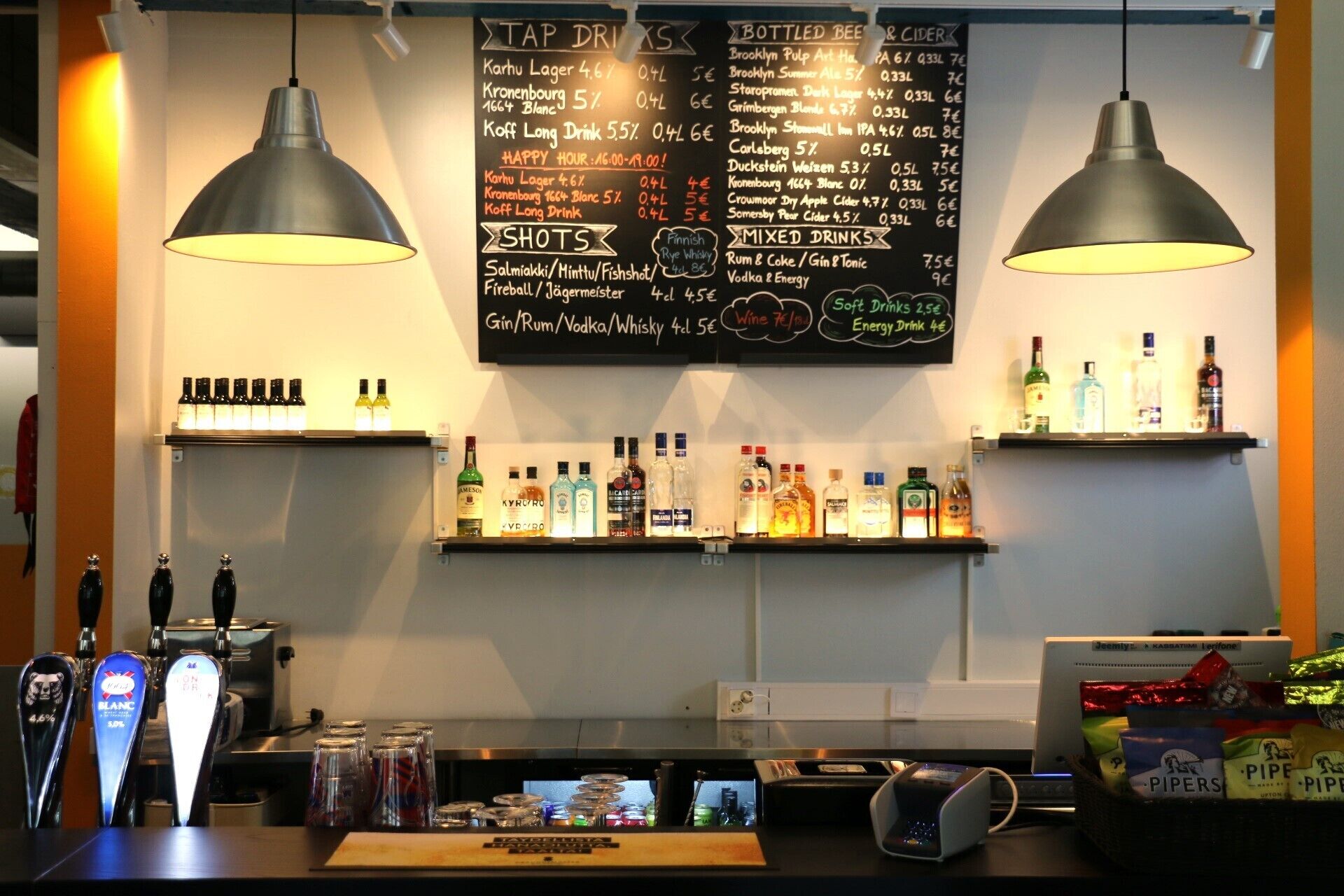 Zona de bar con mostrador de grifos, estanterías llenas de bebidas embotelladas y una pizarra con el menú de bebidas y precios.