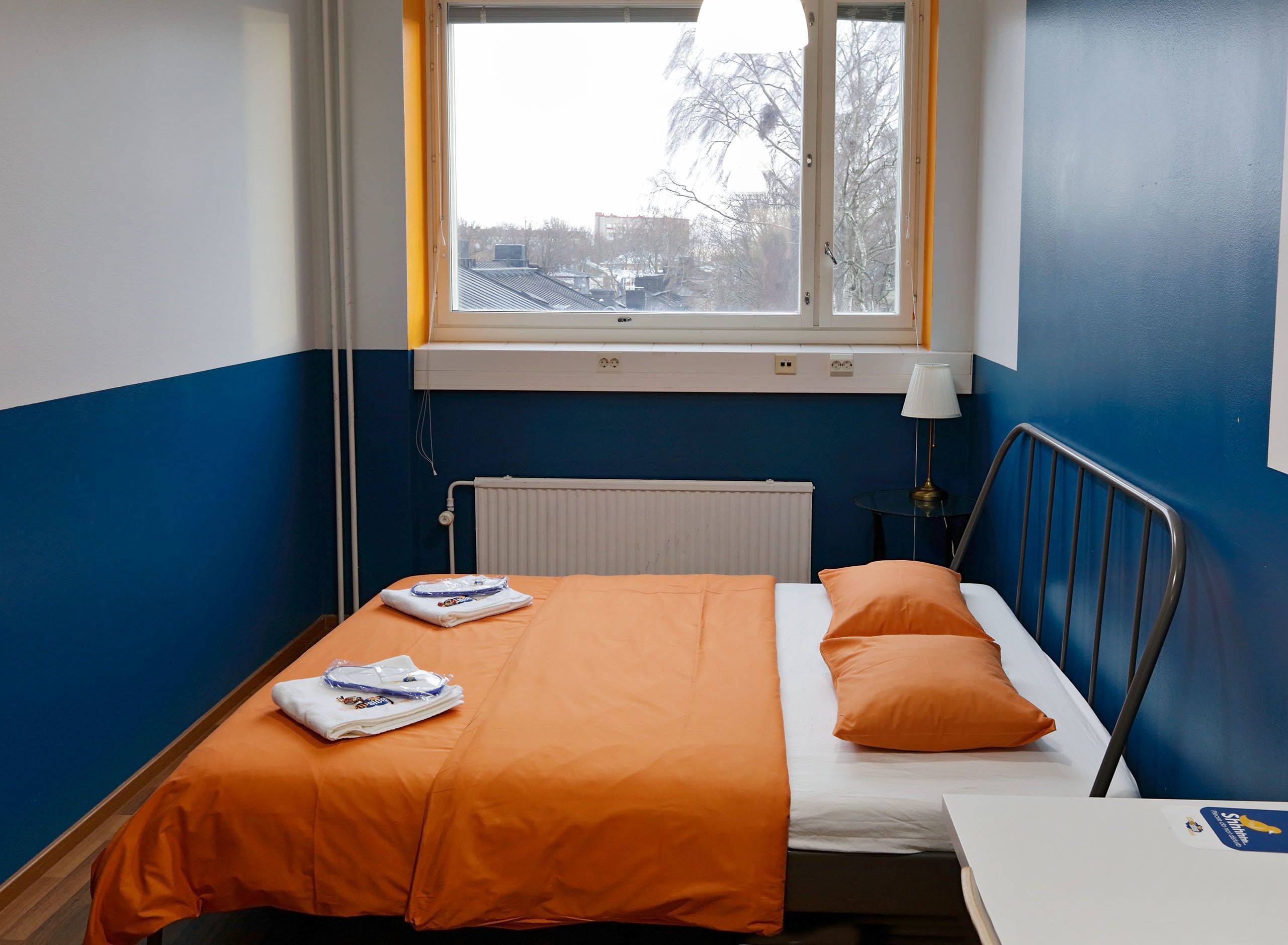 Una cama individual con una colcha y almohadas naranjas, una mesita de noche con lámpara, un radiador y una ventana con vistas a edificios y árboles.