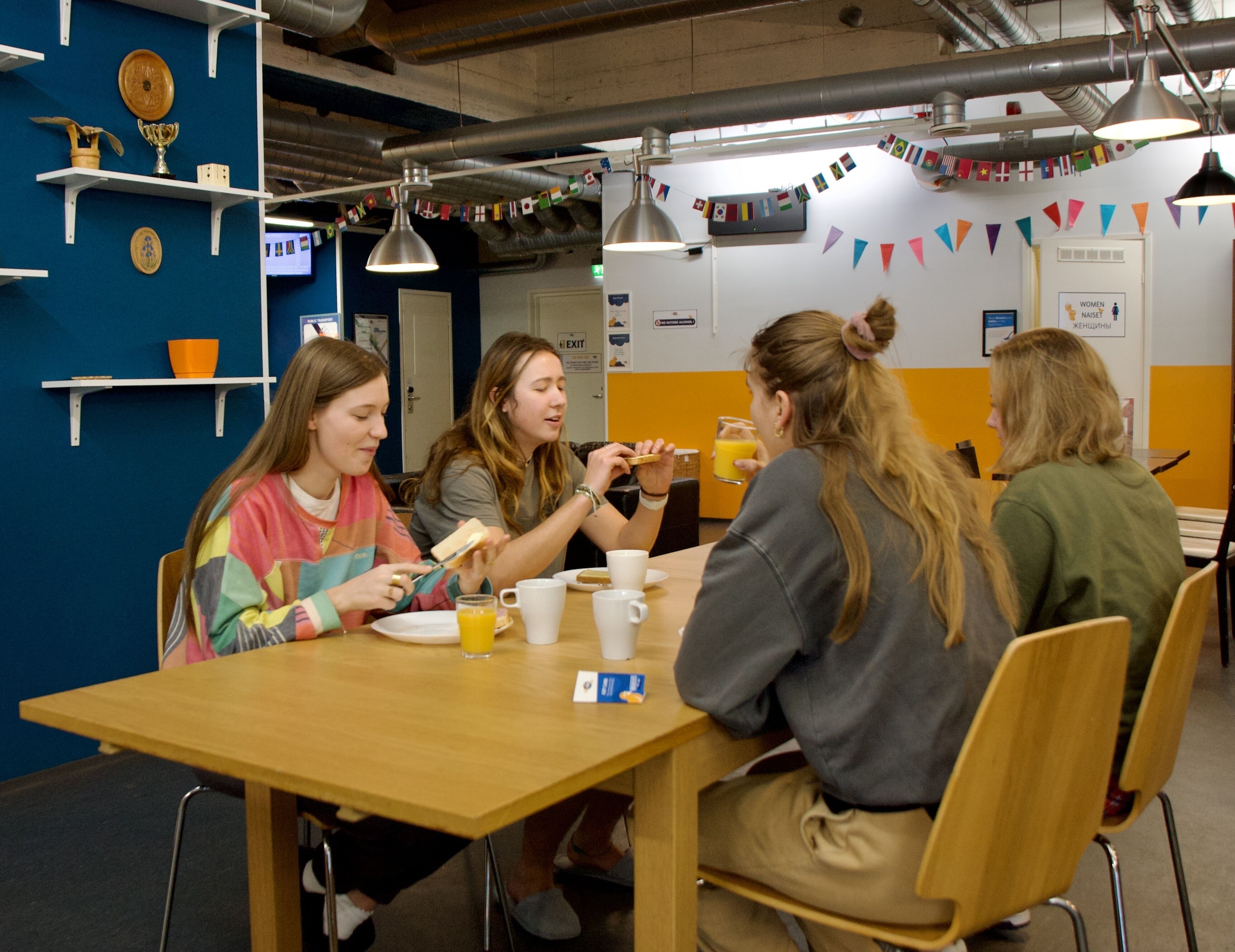 A group of people sitting at a wooden table in a room with blue walls, shelves with various items, and a door with an exit sign.
