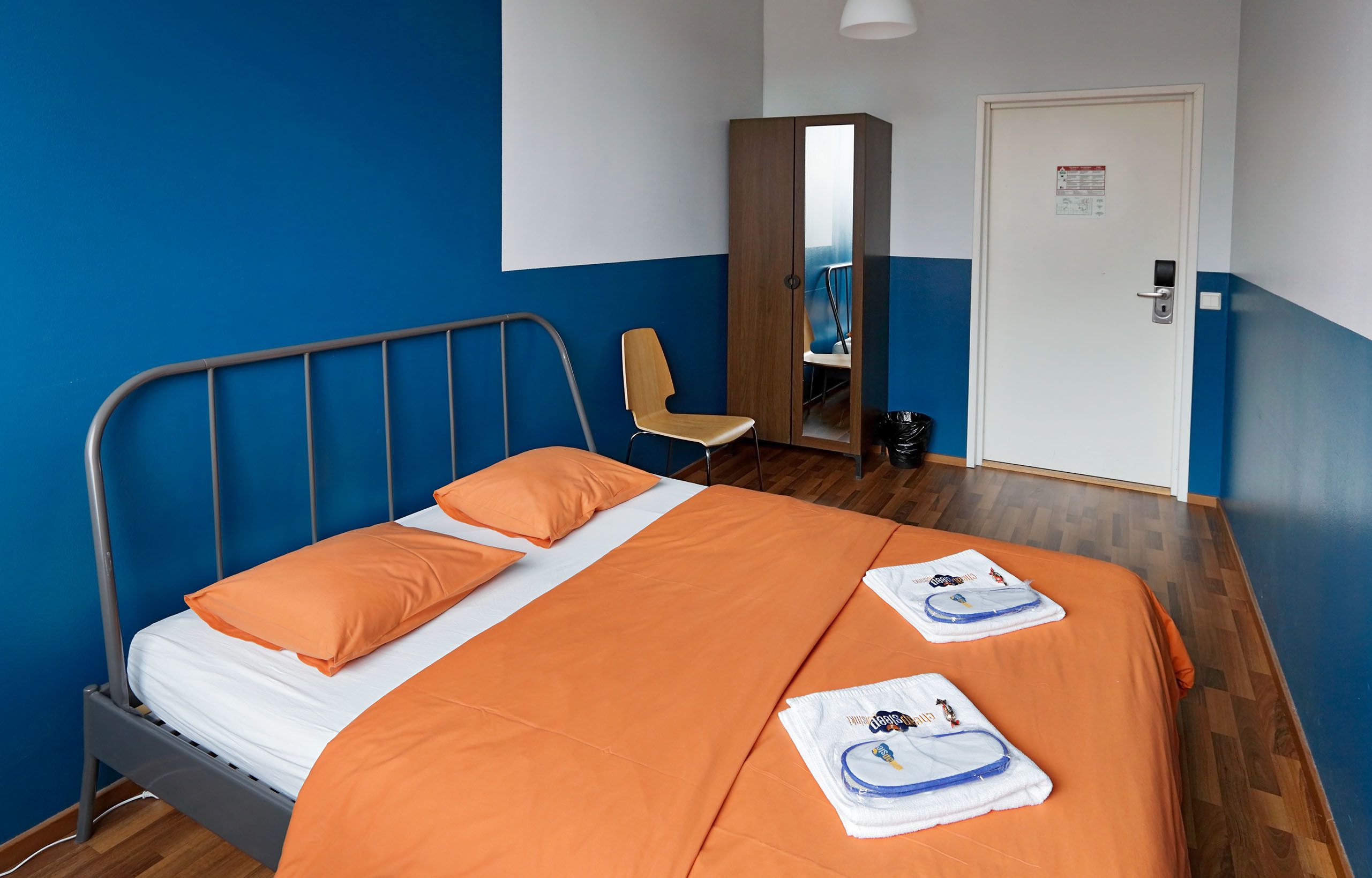 A hotel room with a bed, orange bedding, a wooden wardrobe, a chair, and a door with a sign.