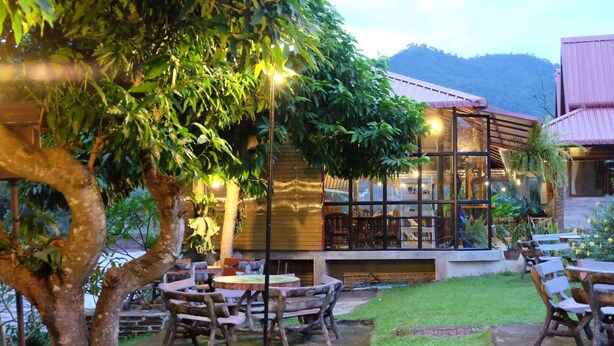 An outdoor seating area with tables and chairs, surrounded by greenery and a building with large windows. - Khun Mai Baan Suan Resort (Mae Ai)