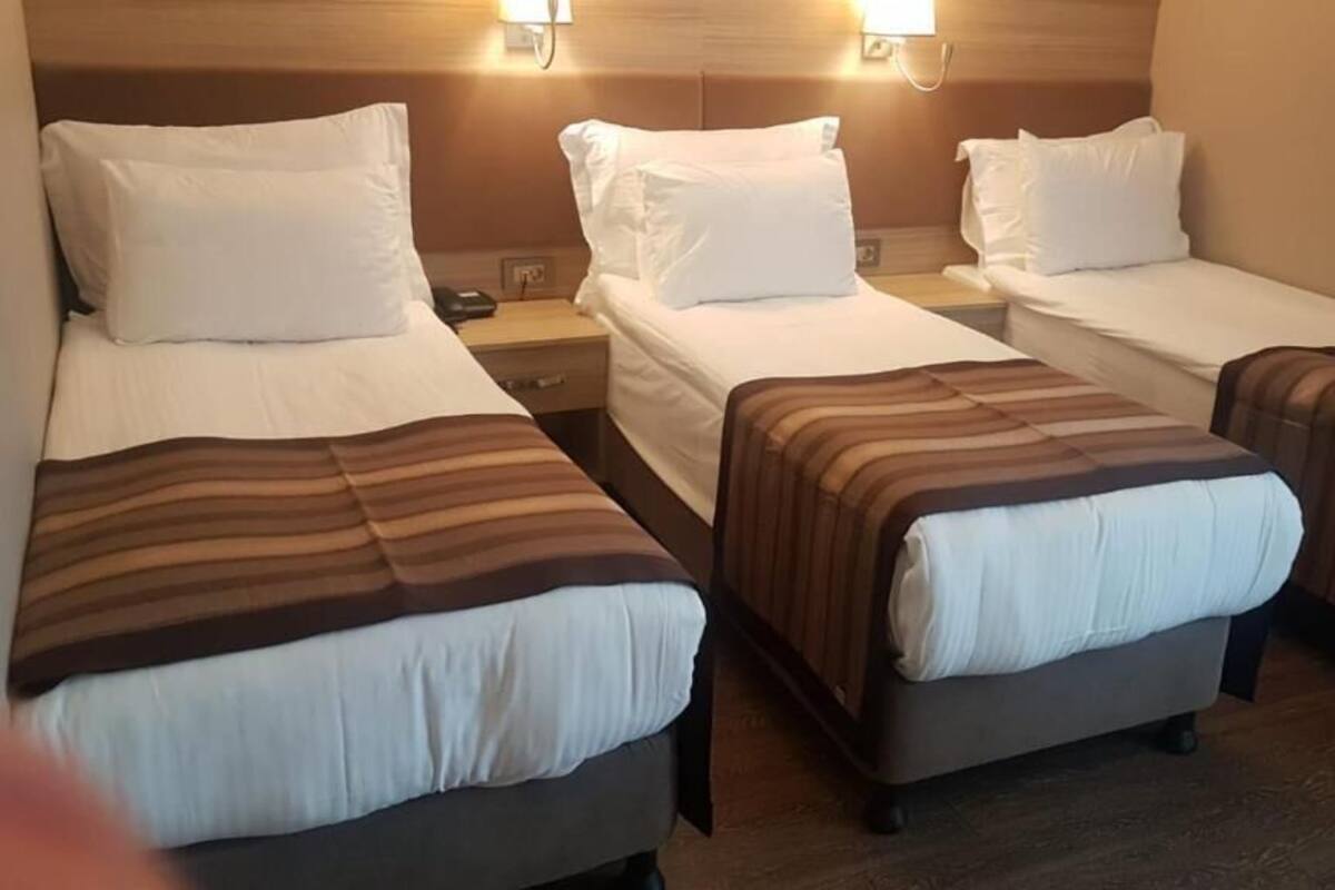 A hotel room with three single beds, each with a striped blanket and white pillows. There are bedside tables with lamps and a nightstand with a phone.