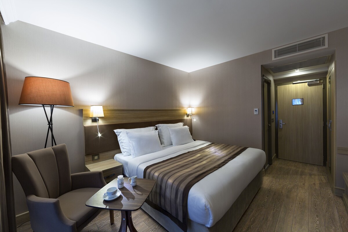 Habitación de hotel con cama, una silla, una mesa y dos lámparas fijadas a la pared.