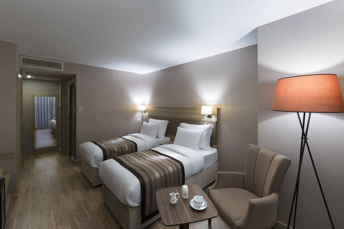 A hotel room with two beds, a chair, a small table with a cup and a pitcher, and a floor lamp.