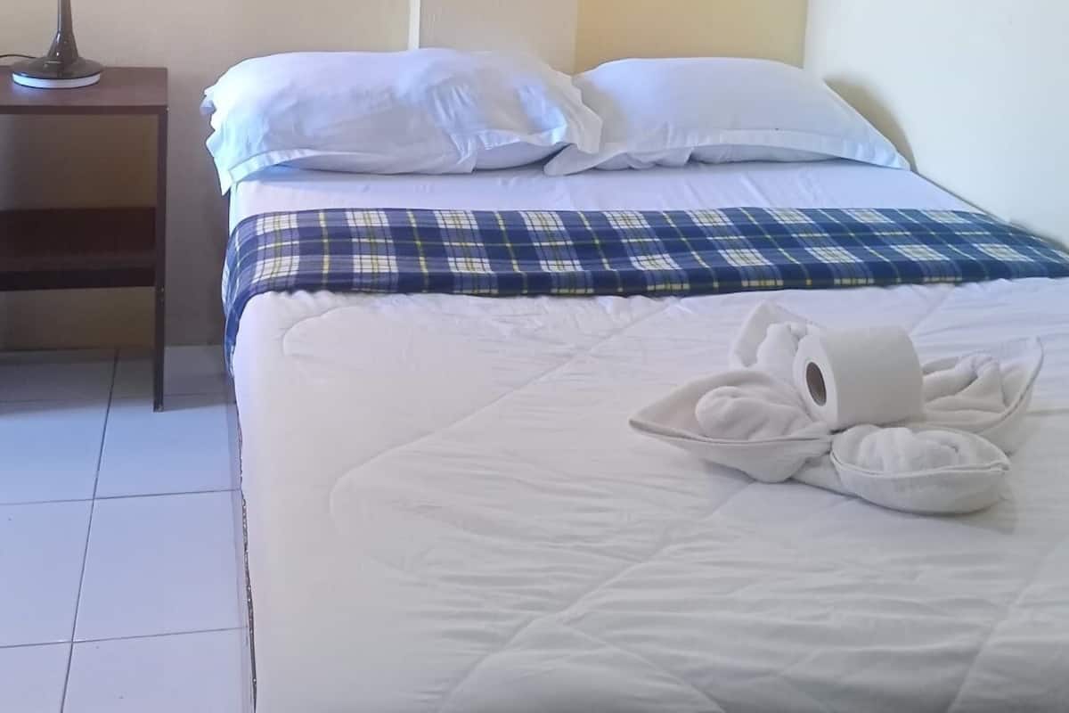 Un lit bien fait, avec une literie blanche et bleue, une lampe de chevet et un rouleau de papier toilette posé sur le sol.