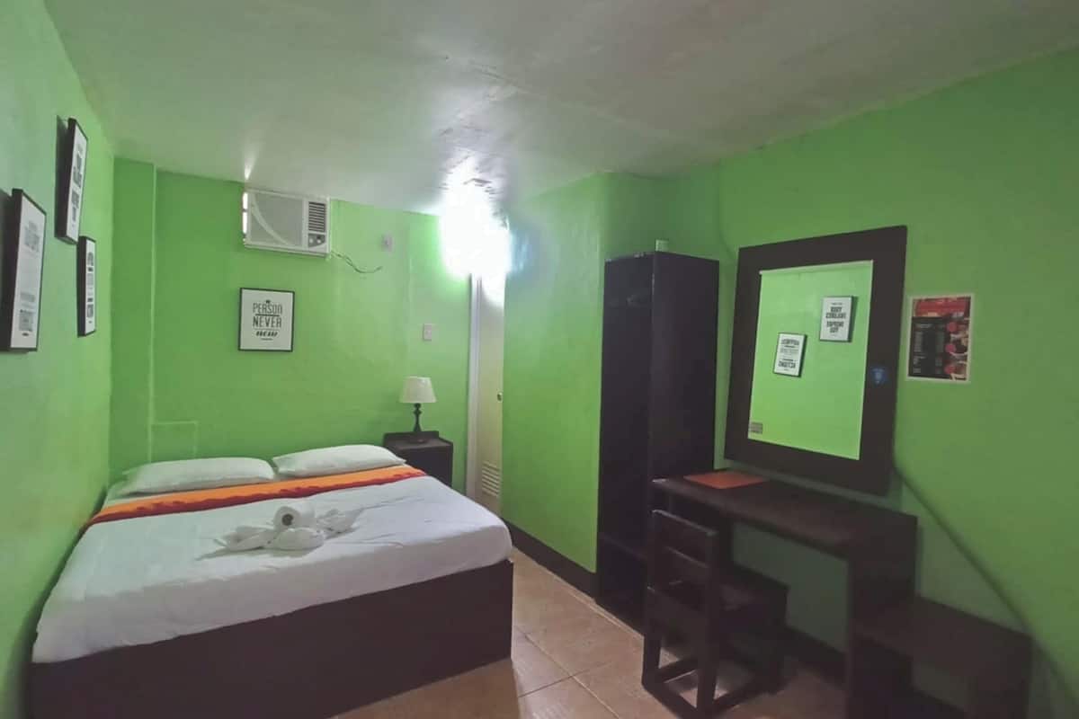 Une pièce aux murs verts, un lit recouvert d’une couverture blanche et orange, un bureau avec une chaise et un miroir accroché au mur.