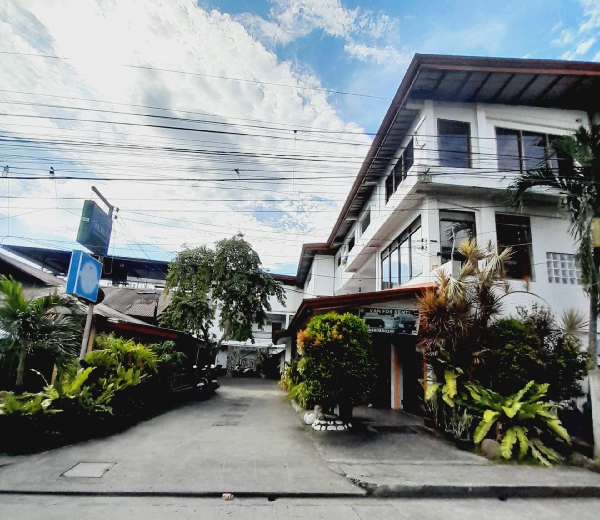 A street view with a van for rent sign, a building with a white facade, and lush greenery.
